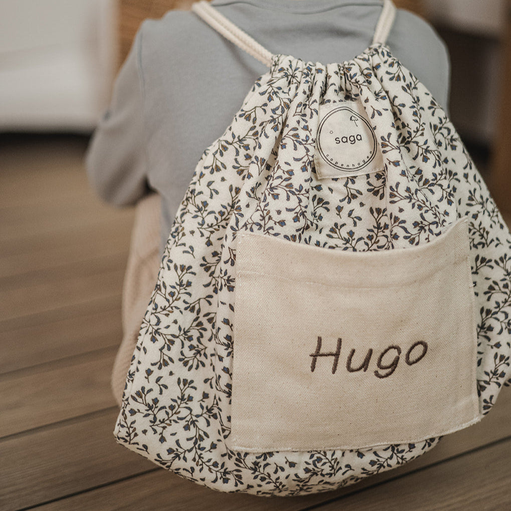 Child wearing a floral drawstring bag with 'Hugo' on a wooden floor